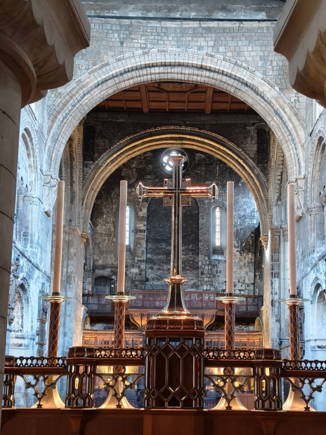 A view from behind the high altar of St Bartholomew the Great, looking through a series of massive Norman stone arches. In the foreground, a polished silver altar cross stands between four tall, slender candles on an ornate wooden screen. The background reveals the weathered, ancient stone walls of the sanctuary, illuminated by soft natural light from high windows, highlighting the church's medieval Romanesque architecture.